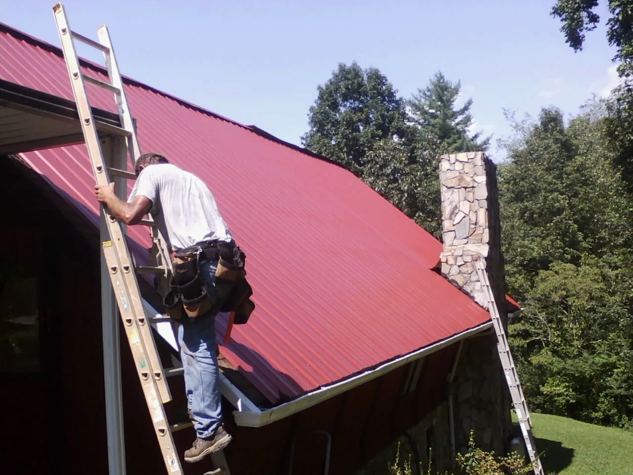 Roofer on a ladder beside a red metal roof and stone chimney — Soffit & Fascia Repair by Roofing Brothers in Bonsall